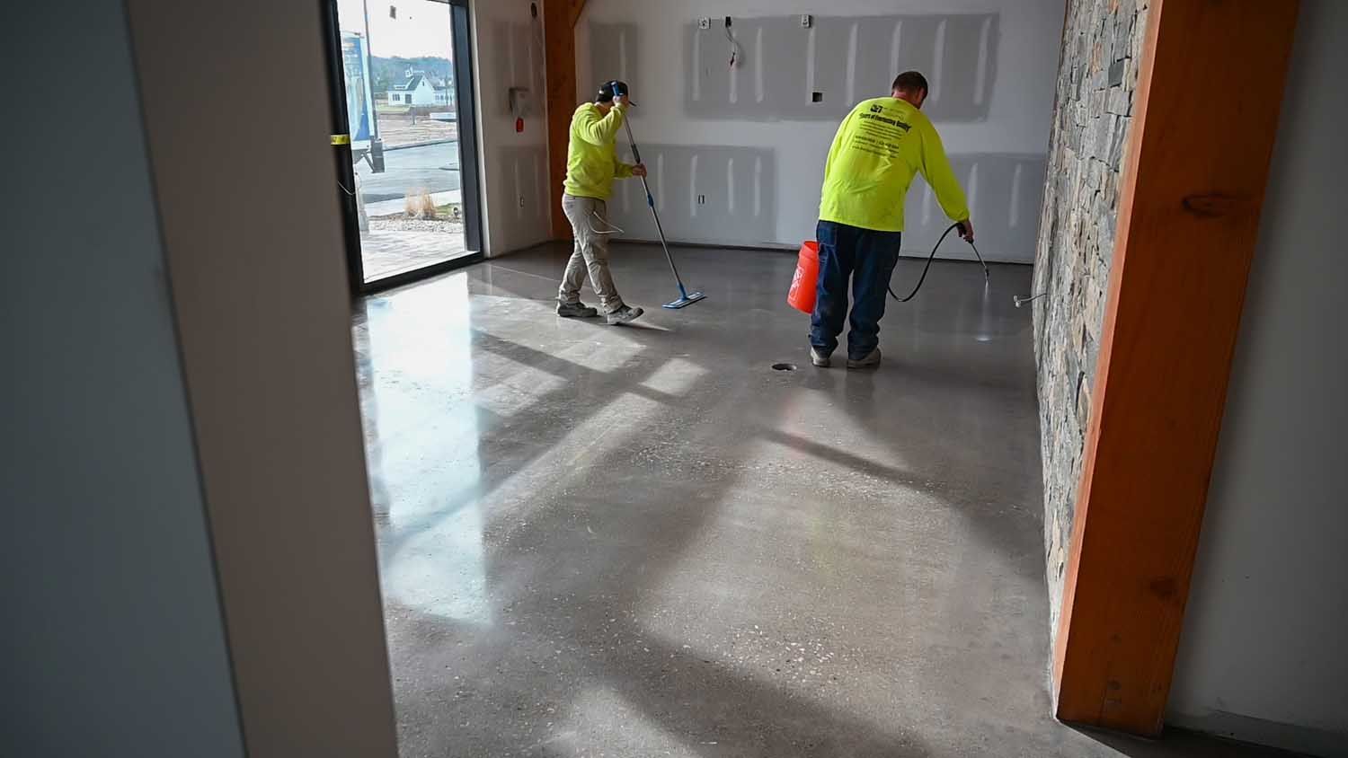 Two workers in yellow jackets mop a bare room floor near a doorway.