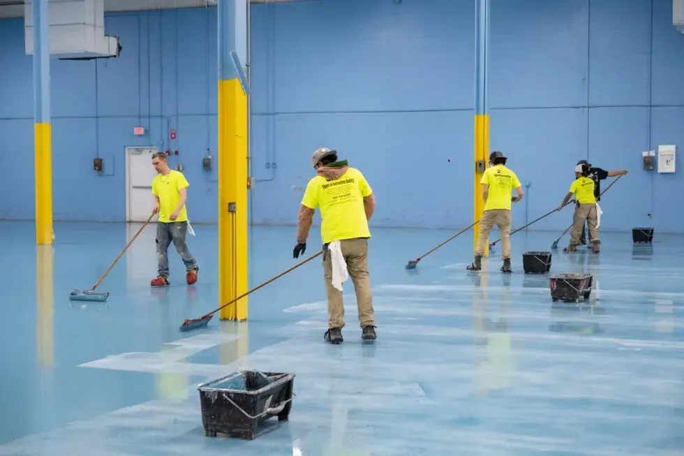 Four workers in yellow shirts use rollers to apply anti-slip epoxy floor coating to a large