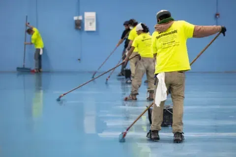 Workers in neon yellow shirts painting a glossy blue floor with long-handled rollers