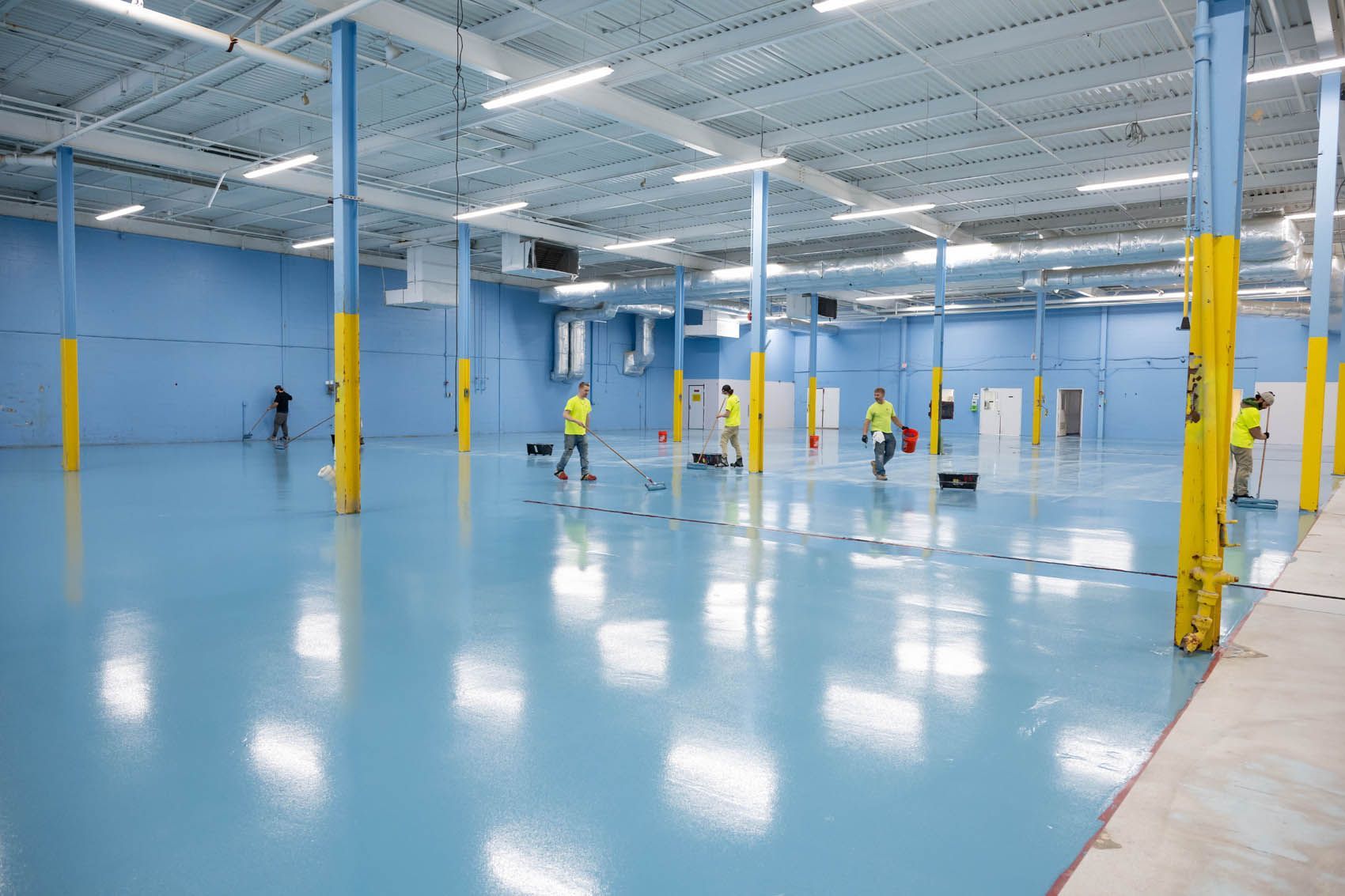 Large empty blue-floored warehouse with a few workers in yellow safety vests and equipment