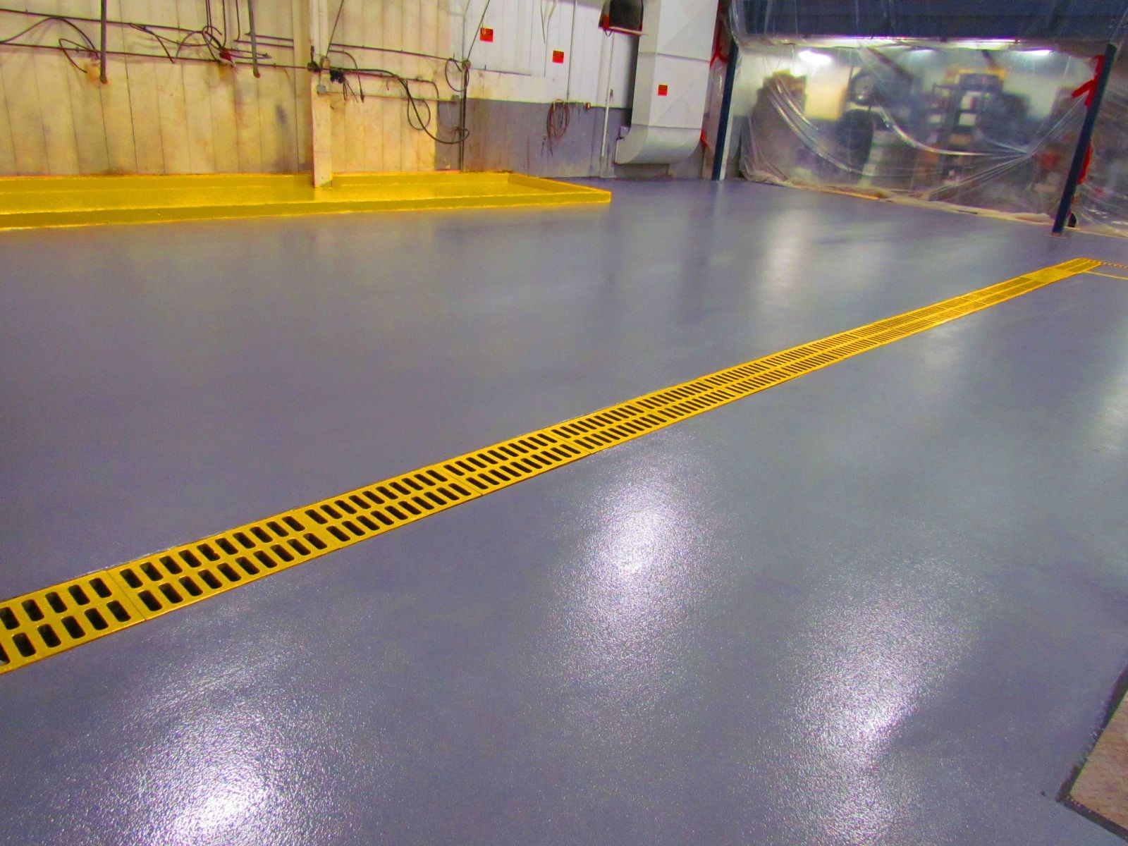 Industrial floor with a yellow grated drain trench across a gray epoxy surface in a warehouse.