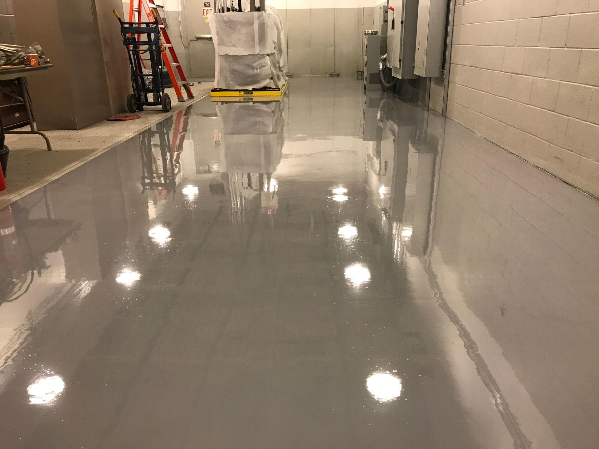 Shiny gray floor in a hallway, reflecting lights, with cleaning equipment and covered items at the far end.