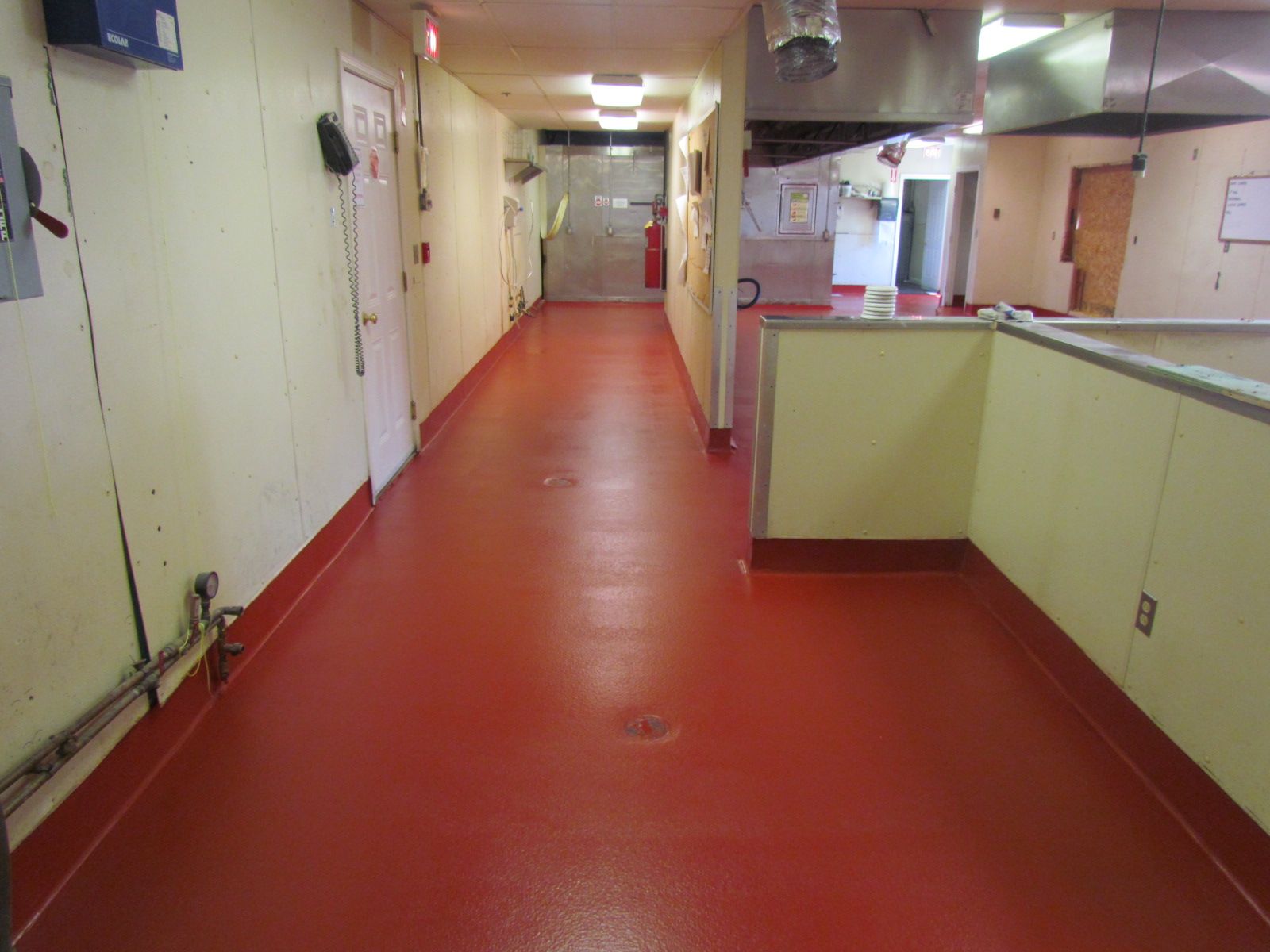 Narrow indoor hallway with a red floor and cream-colored walls, leading to a brighter area ahead.