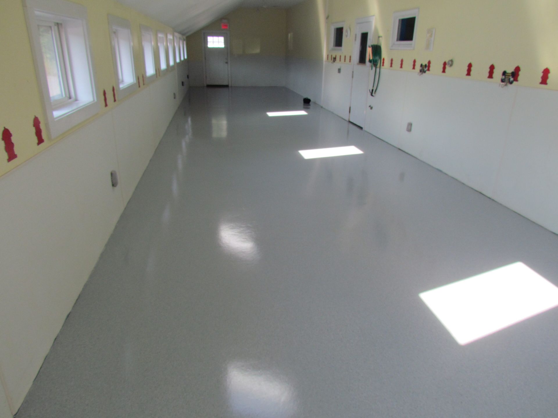 Empty bright hallway with white walls, windows on the left, and sunlight patches on the floor.