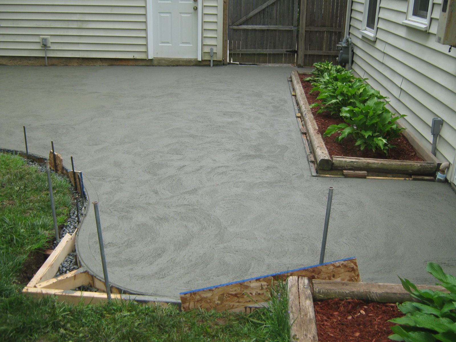 Freshly poured concrete patio in a backyard beside a house, bordered by wooden forms and garden beds