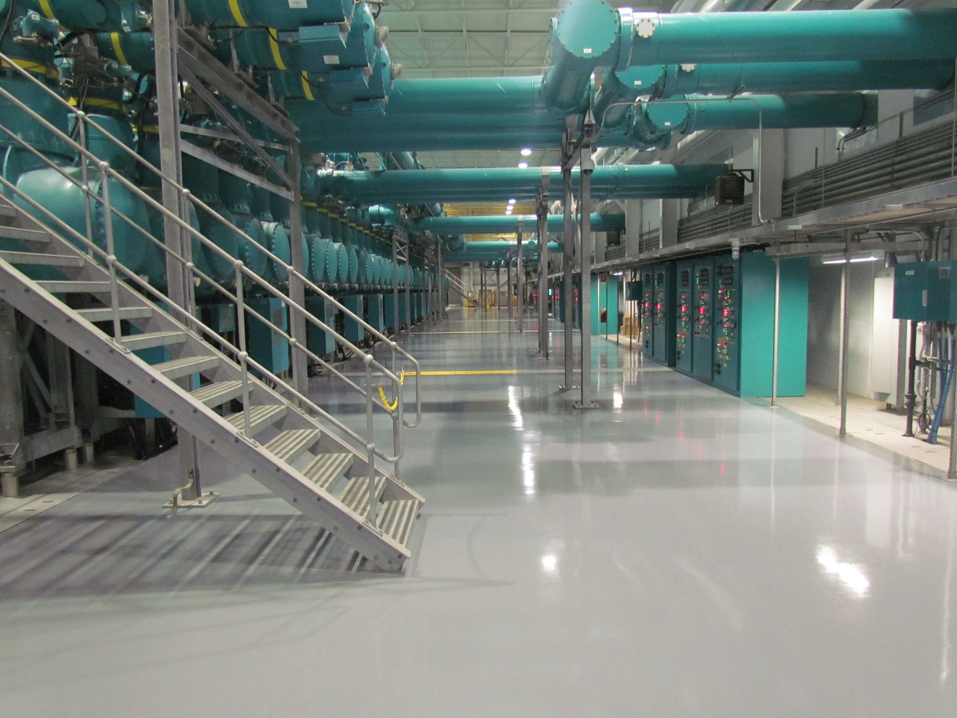 Industrial facility interior with teal pipes, metal stairs, and a polished concrete floor