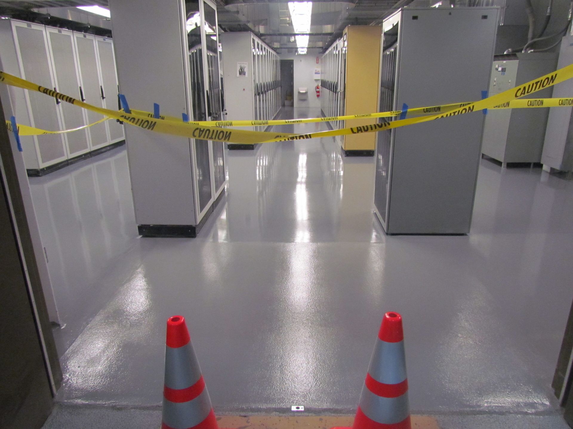 Caution tape blocks a bright hallway with two orange cones in the foreground.