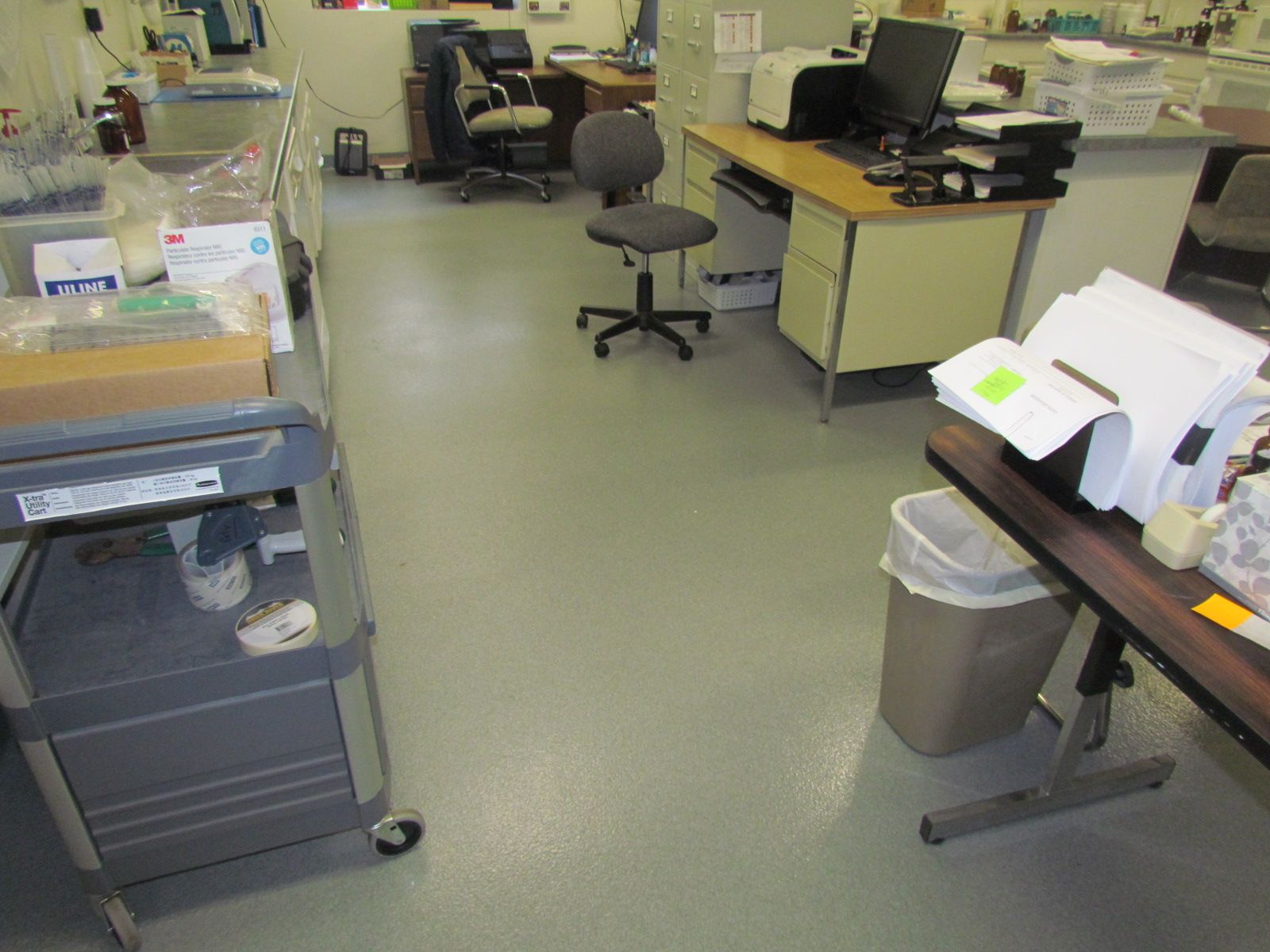 Cluttered office workspace with desks, computers, rolling chairs, and a trash bin on a green floor