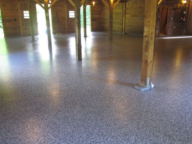 Empty wooden hall with polished speckled floor and support posts, lit by daylight from windows.