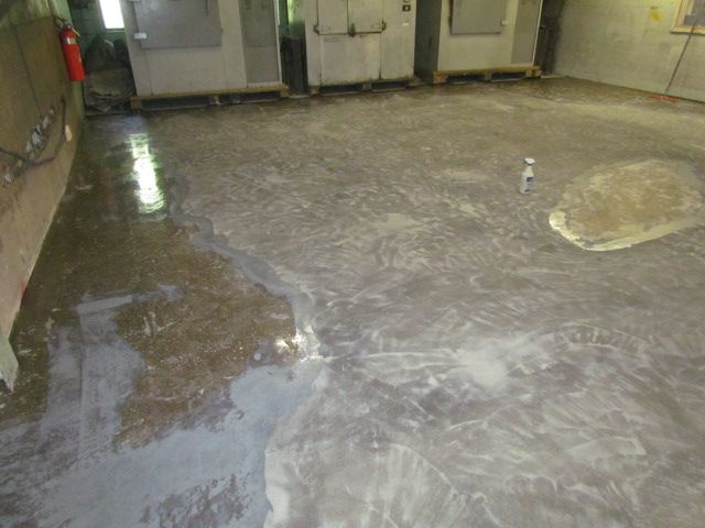 Wet, reflective concrete floor in an empty industrial room with doors and equipment along the walls