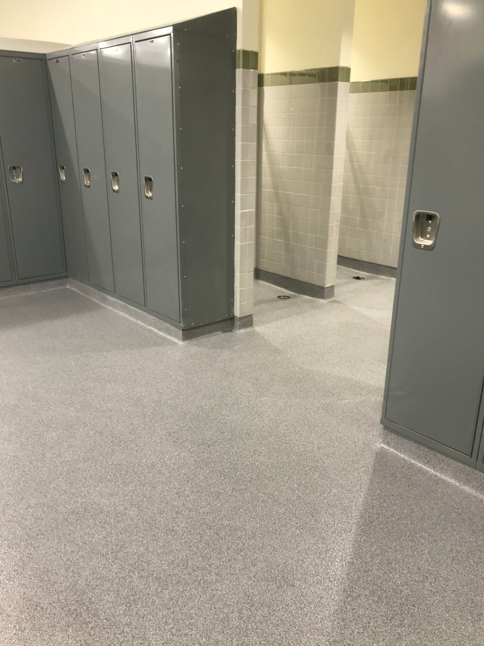 Empty school hallway with gray lockers and restroom stalls on a speckled tile floor