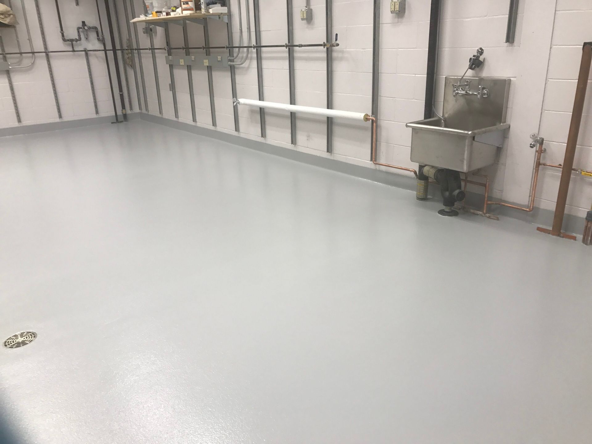 Empty industrial room with smooth gray floor, white walls, pipes, and a utility sink.