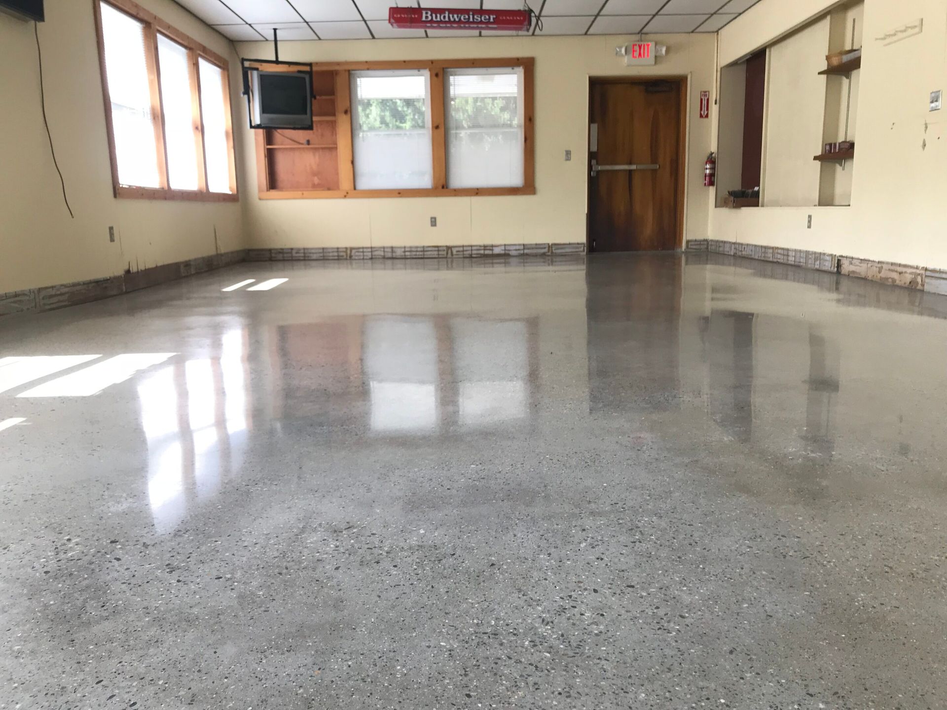 Empty indoor hall with polished concrete floor, windows, wall shelves, and a wooden door at the far end.