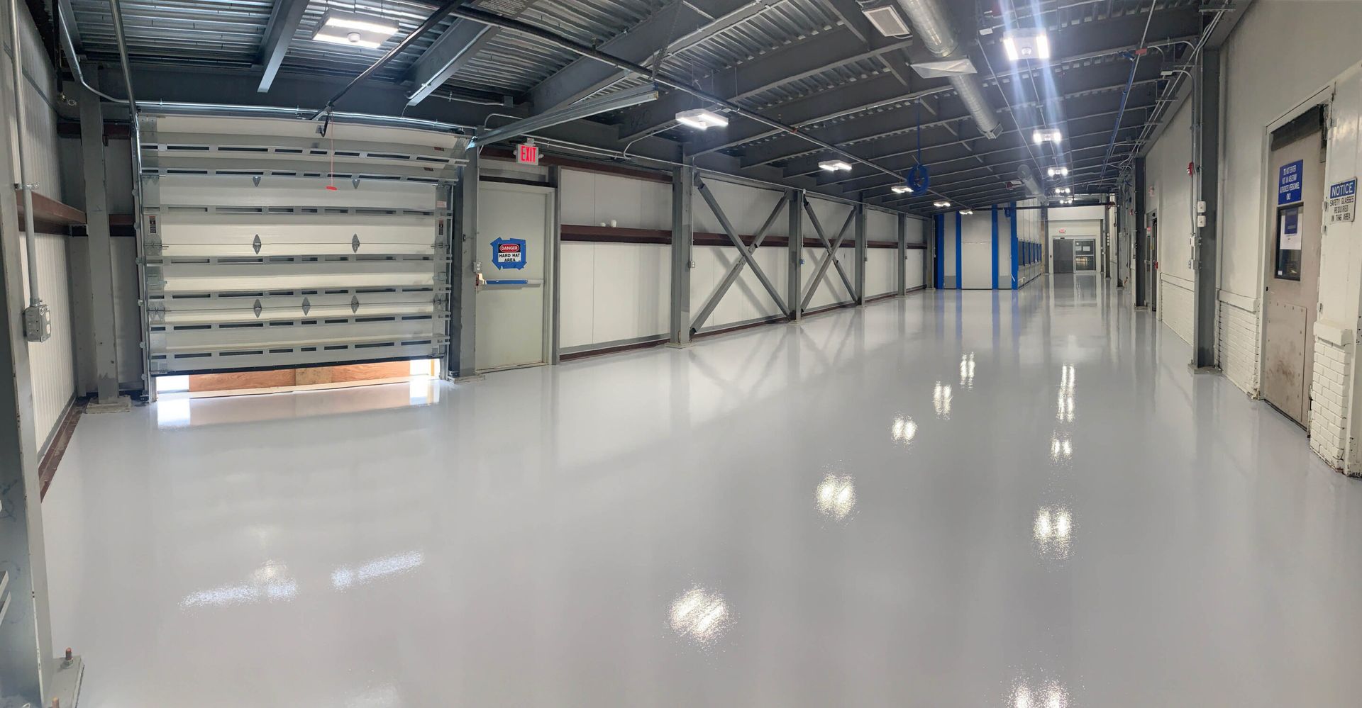 Empty industrial warehouse interior with glossy white floor, overhead lights, and large loading door