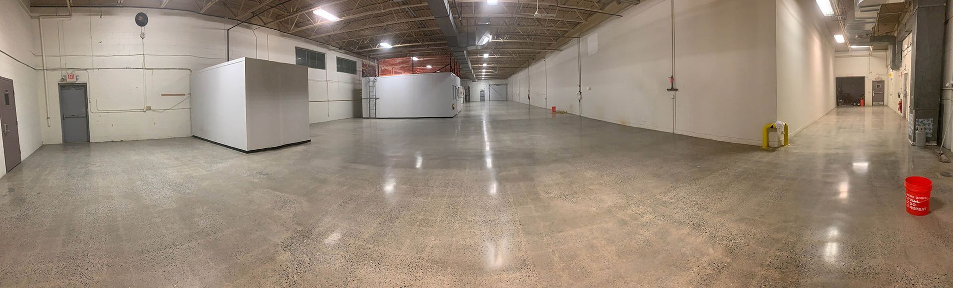 Empty industrial warehouse with concrete floor, white walls, overhead lights, and a red bucket near the right wall.