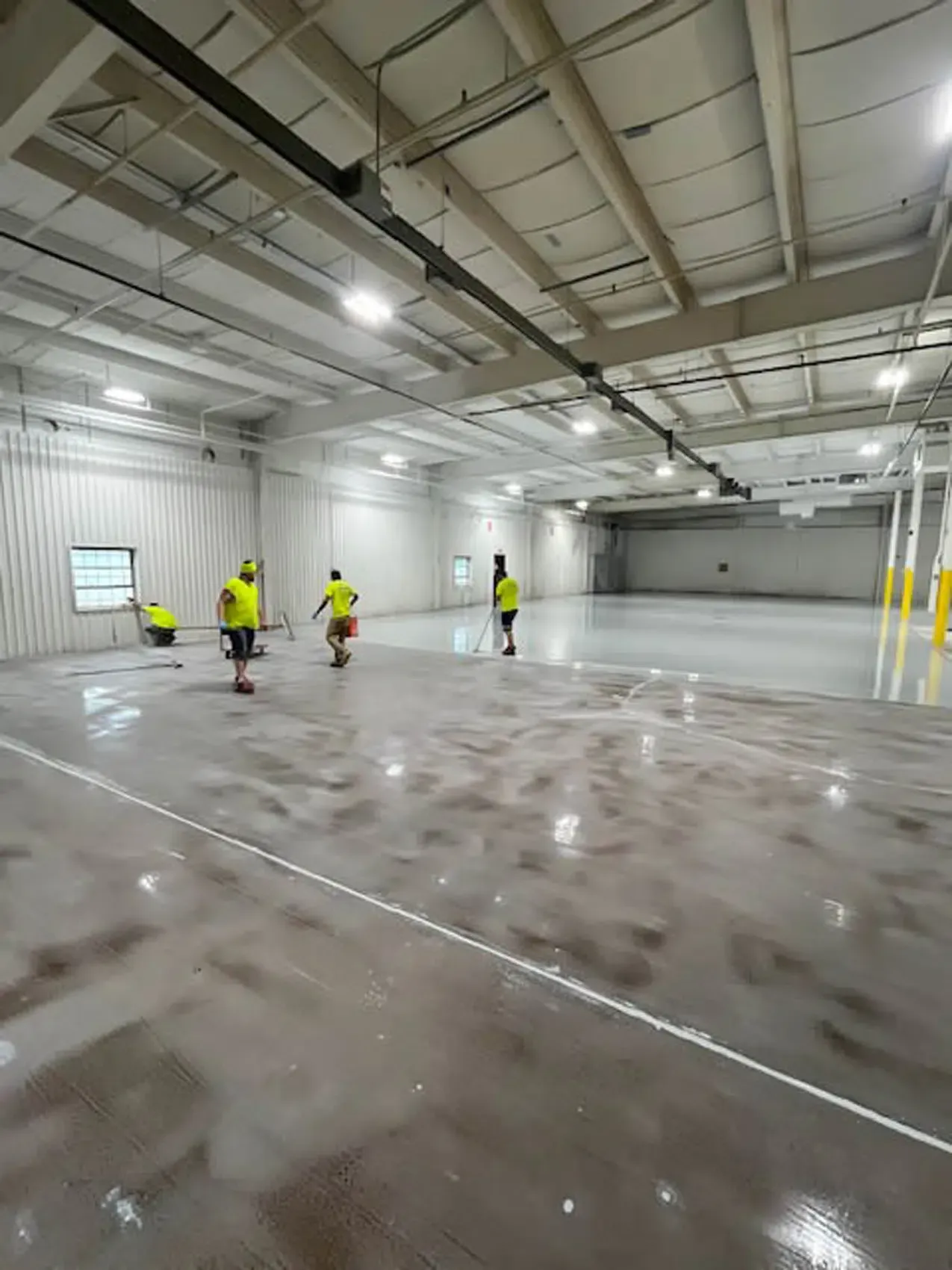 Workers in yellow vests inspect a large empty industrial warehouse floor.