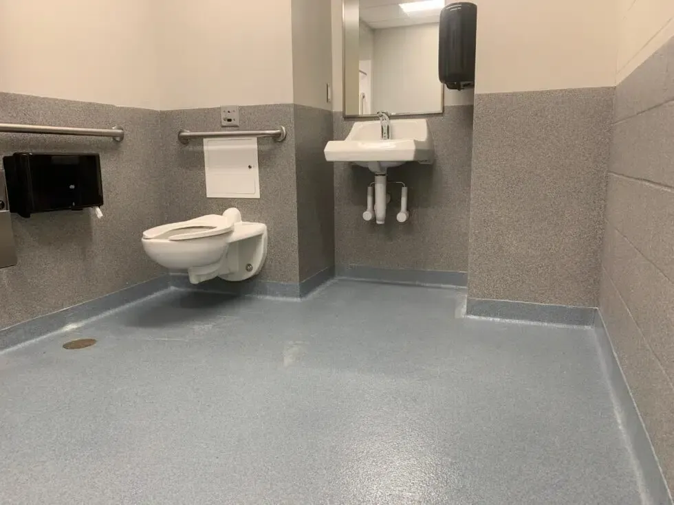 Accessible restroom with wall-mounted toilet and sink, grab bars, and blue-gray floor.