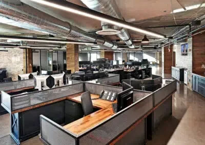 Open-plan office with desks, monitors, and exposed industrial ceiling and brick walls