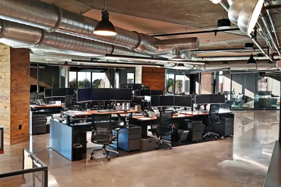 Open-plan modern office with desks, computer monitors, and exposed brick and ductwork ceiling
