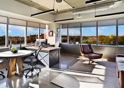 Bright modern office lounge with large windows, desks, chairs, and sunlight streaming across the floor
