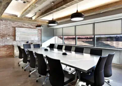 Modern conference room with long white table, black chairs, and large windows exposing brick walls