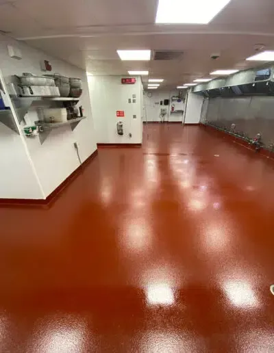 Empty commercial kitchen with red epoxy floor, stainless counters, and shelves along the walls.