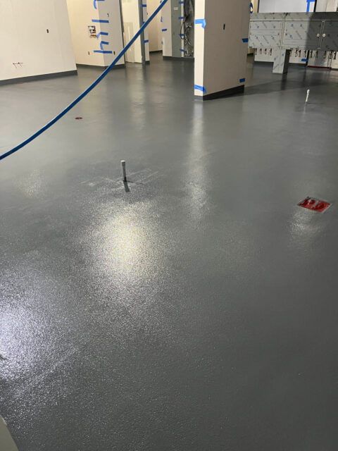 Empty indoor room with gray floor, blue hose, and exposed floor fixtures near pillars.