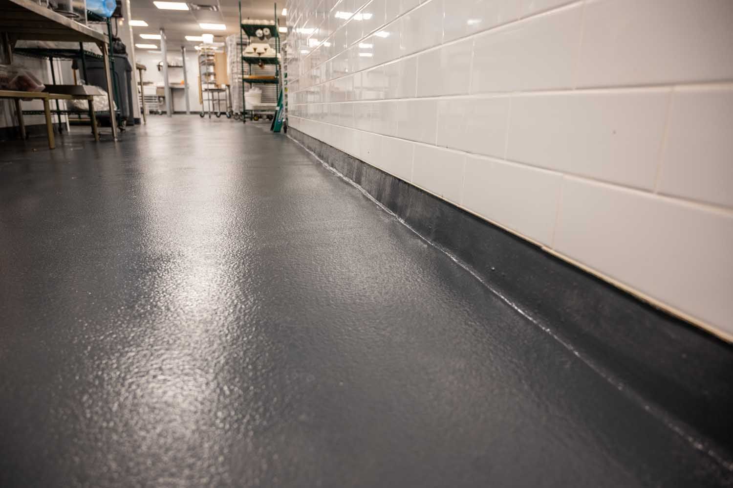 Low-angle view of a clean kitchen floor beside white tiled wall and stainless steel equipment