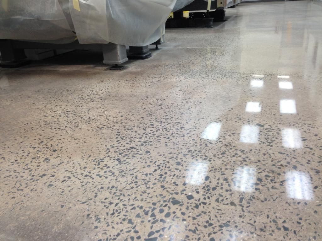 Polished speckled floor in a warehouse aisle with covered equipment and bright window reflections