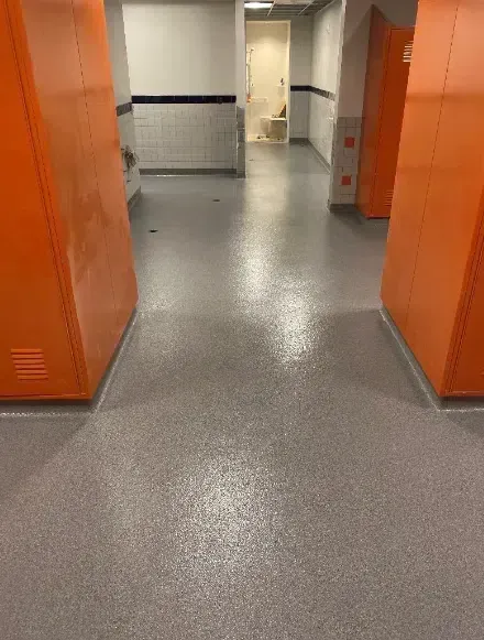 Hallway with orange lockers on both sides and a gray speckled floor, leading to a doorway ahead