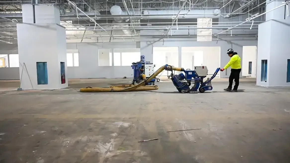 Worker in neon jacket using a floor grinding machine in a large unfinished white warehouse.