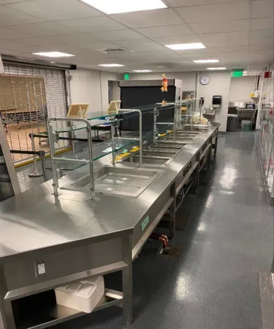 Empty cafeteria serving line with stainless steel counters and tiled floor under bright ceiling lights