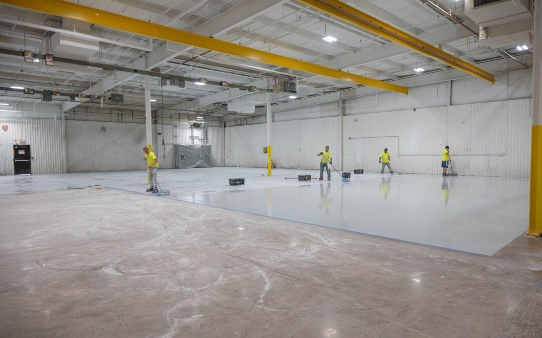 Four workers in yellow shirts are applying a coating or paint to the floor of a large industrial