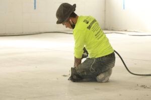 Worker in a neon shirt kneeling and using a power tool on a concrete floor indoors
