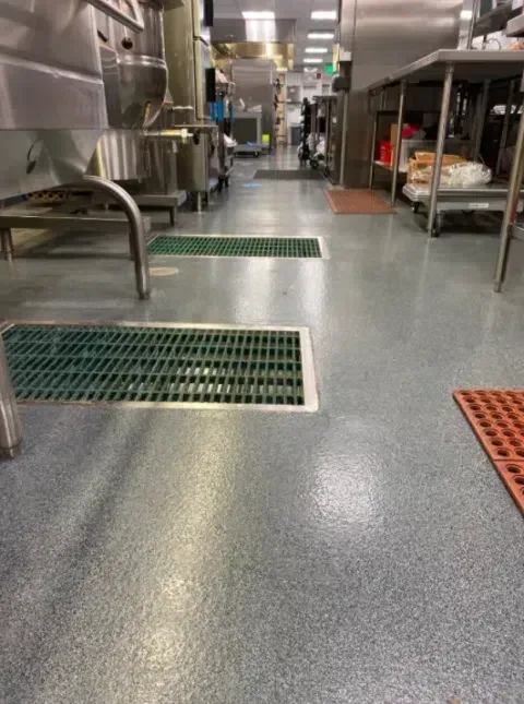 Industrial kitchen aisle with stainless steel equipment and floor drains on a gray floor.