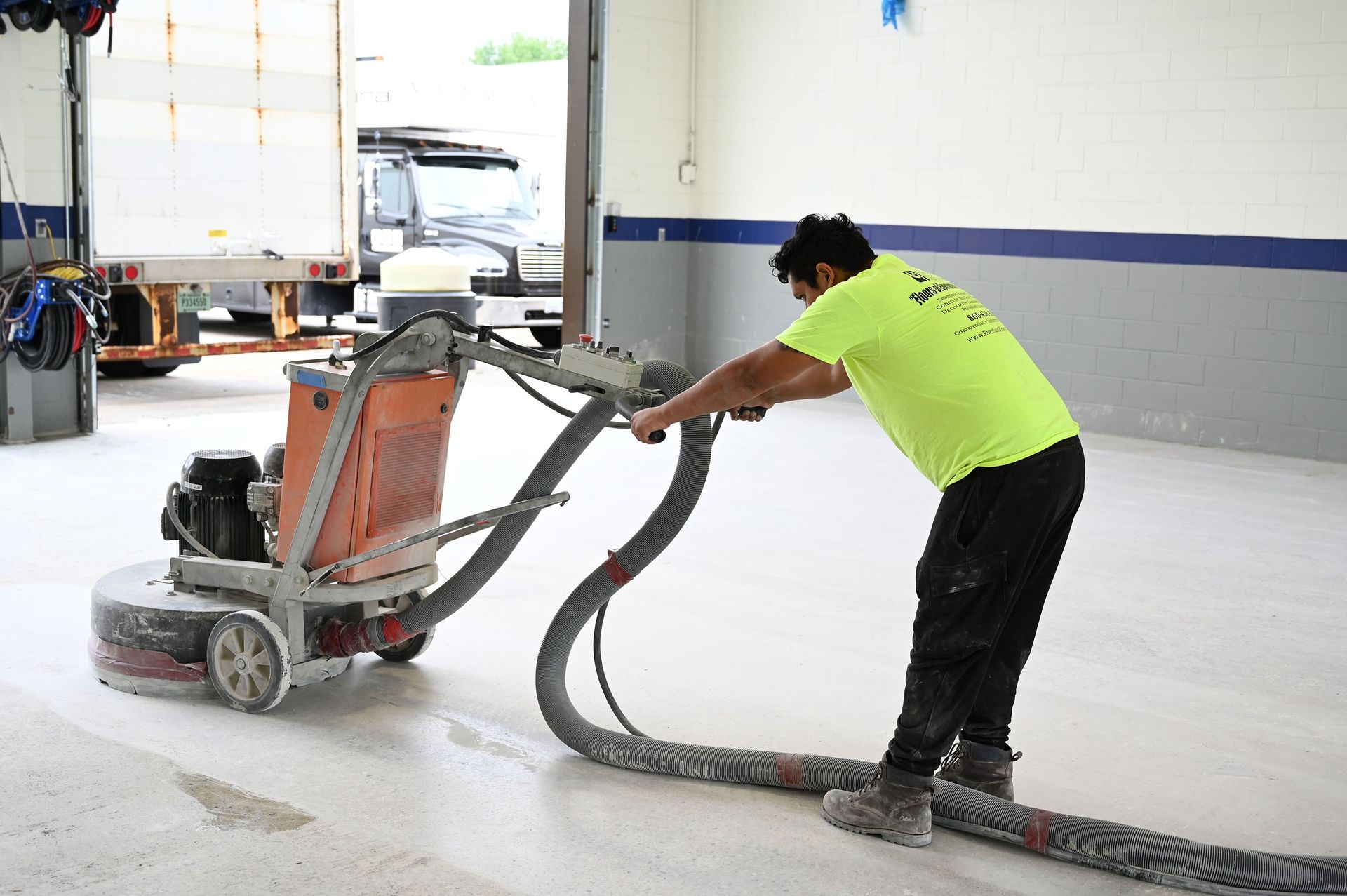 Worker in neon yellow shirt sanding a floor with a floor grinder and hose in a garage workshop.