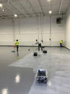 Workers painting a large empty warehouse floor with paint buckets and rollers