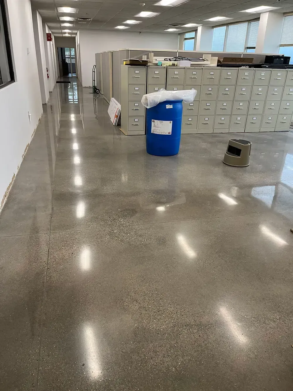 Office hallway with polished floor, cubicles, file cabinets, and a blue recycling bin.