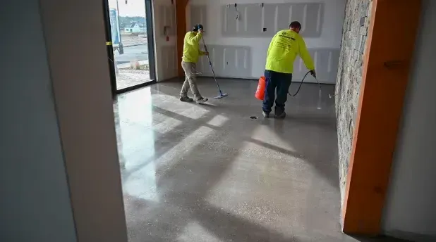 Two workers in yellow shirts mopping a bright empty room with large windows
