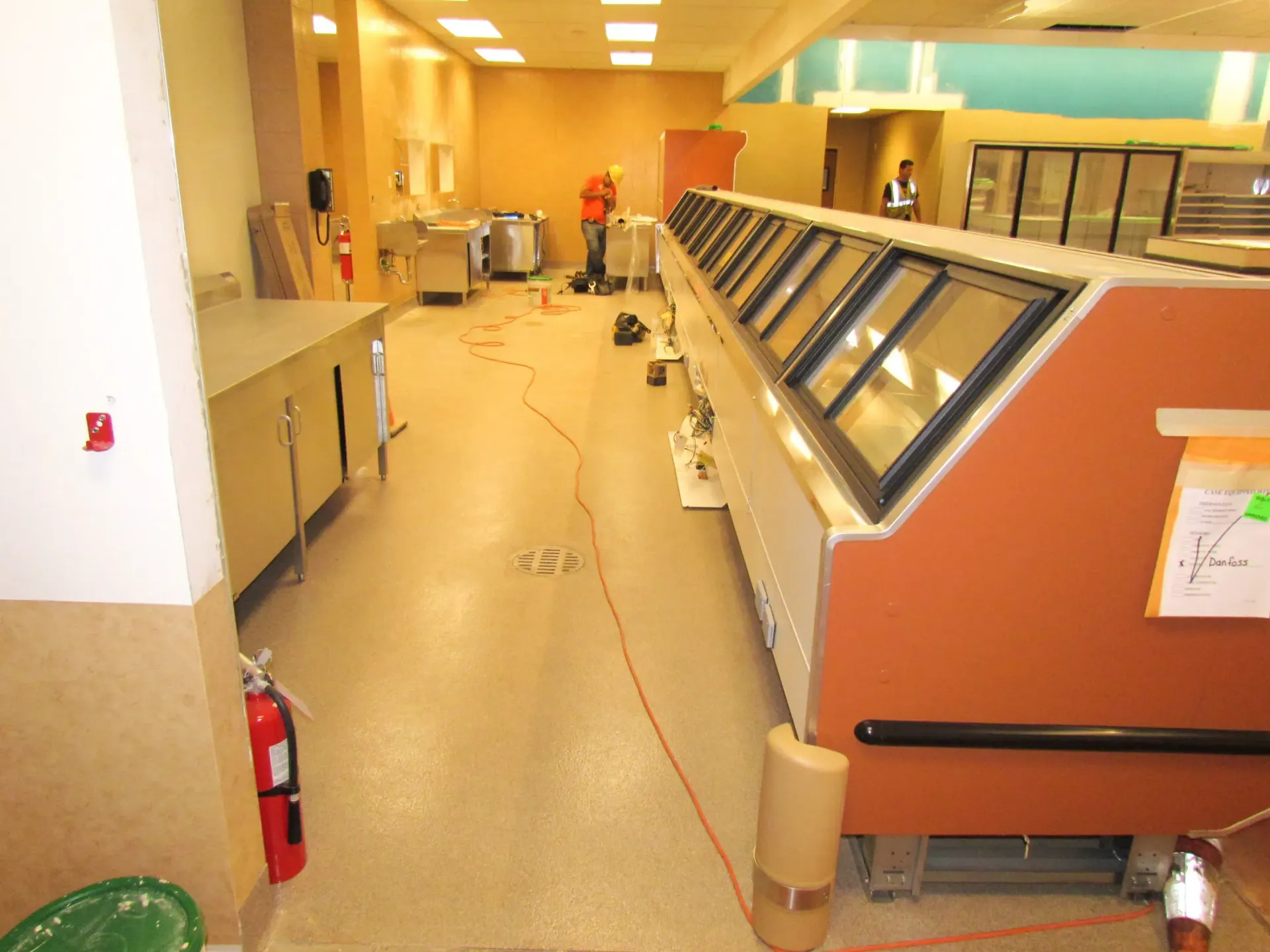Grocery store deli counter with glass display cases and a fire extinguisher in a brightly lit aisle