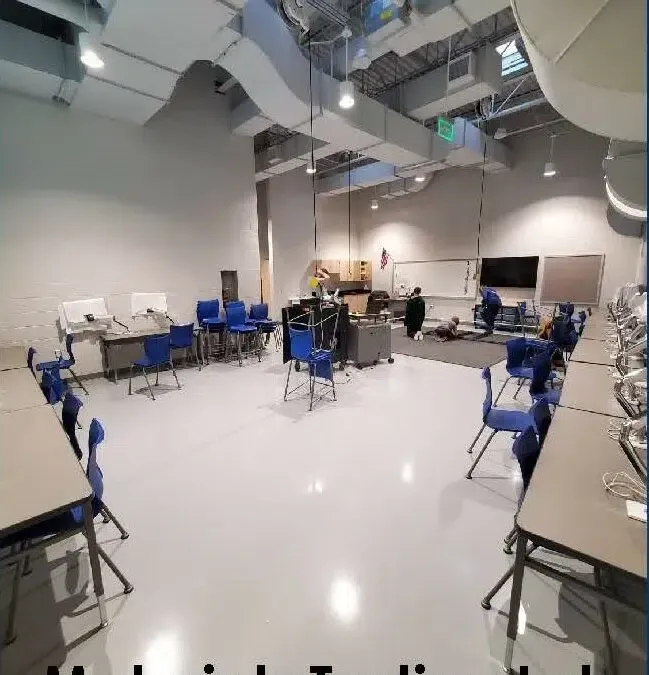 Empty studio classroom with blue chairs, tables, and overhead ductwork; one person near the back.