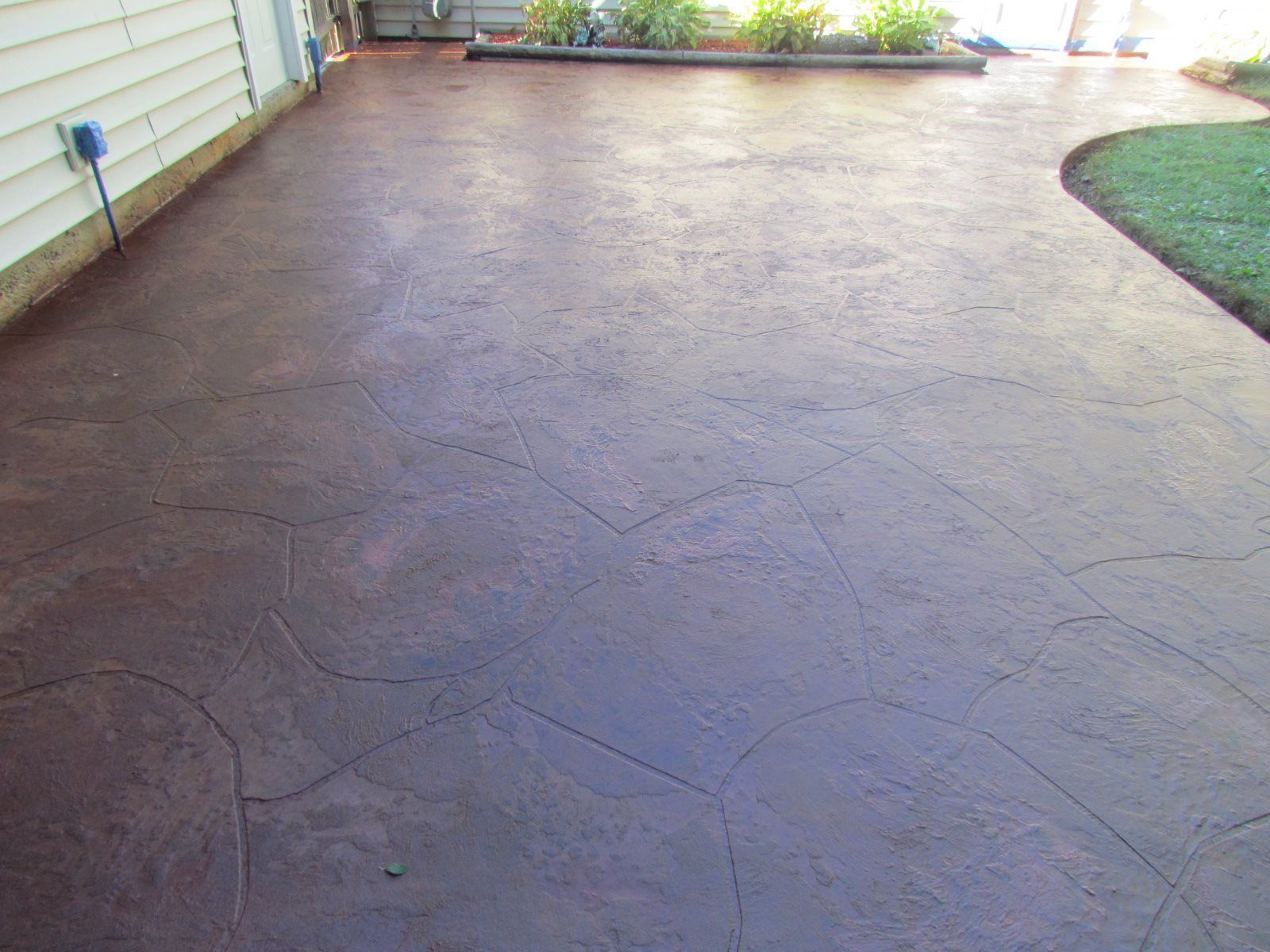 Empty stamped concrete patio beside a house, with a curved grassy edge and flower bed in the distance