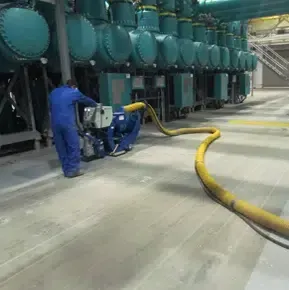 Worker in blue coveralls operating equipment beside large teal industrial tanks on a factory floor.