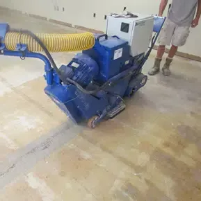 Blue floor grinder with yellow vacuum hose polishing a concrete floor in a workshop