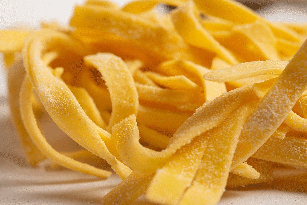 Primo piano di fettuccine crude, di colore giallo chiaro, disposte su una superficie bianca.
