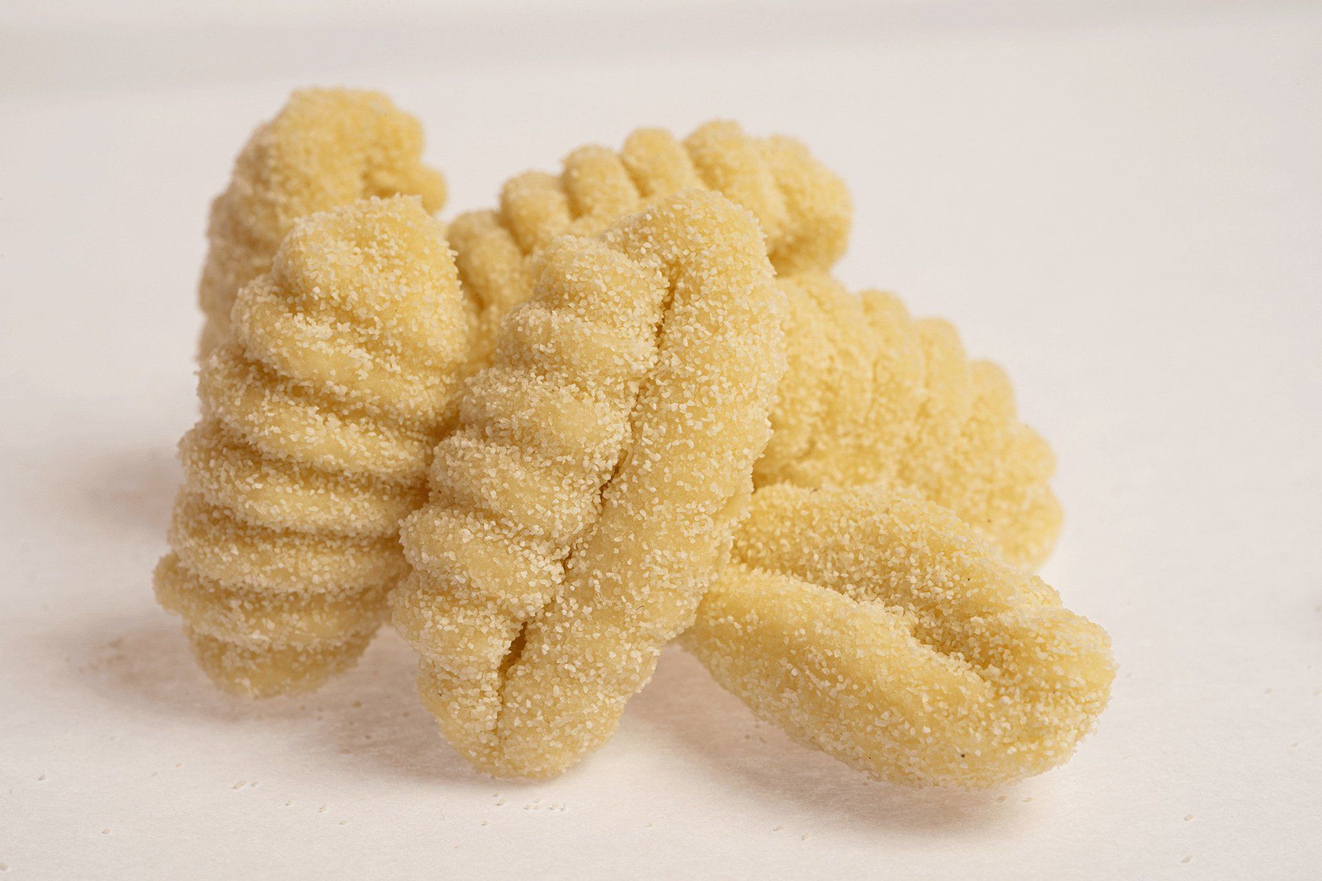Primo piano di quattro formati di pasta rigati, di colore giallo chiaro, probabilmente cavatelli, su sfondo bianco.
