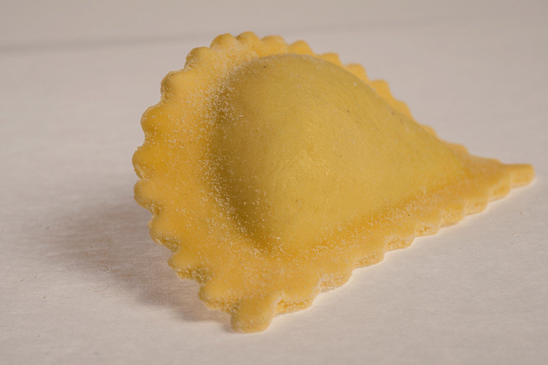 Primo piano di un singolo raviolo crudo, giallo con bordo smerlato, su una superficie bianca.