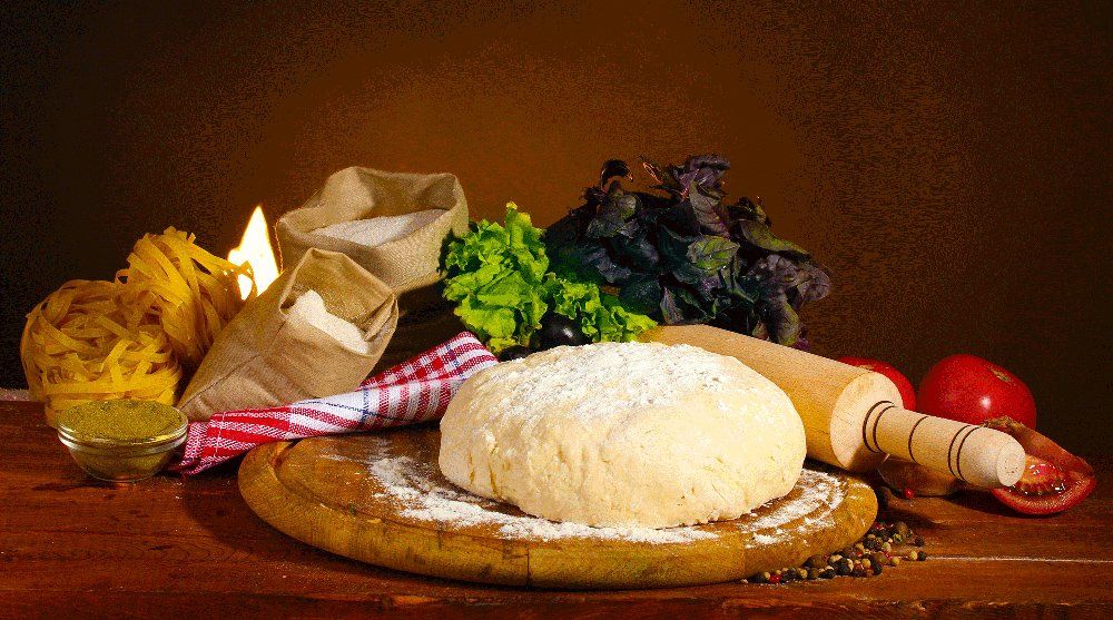 Impasto, farina e ingredienti per la pasta su una superficie di legno, tra cui pomodori, basilico e un mattarello.