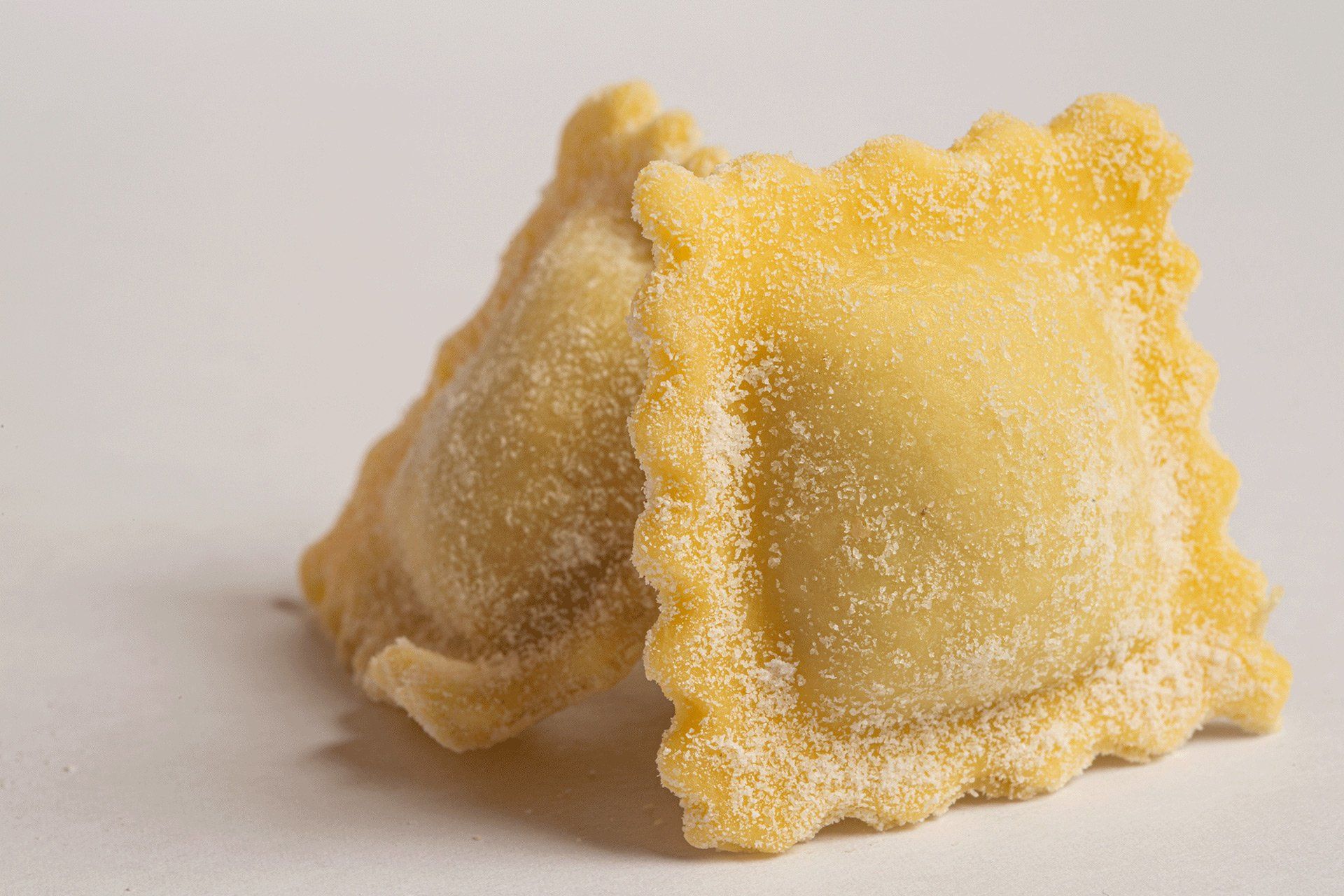 Due ravioli di pasta cruda, di forma quadrata con i bordi arricciati, su una superficie bianca.