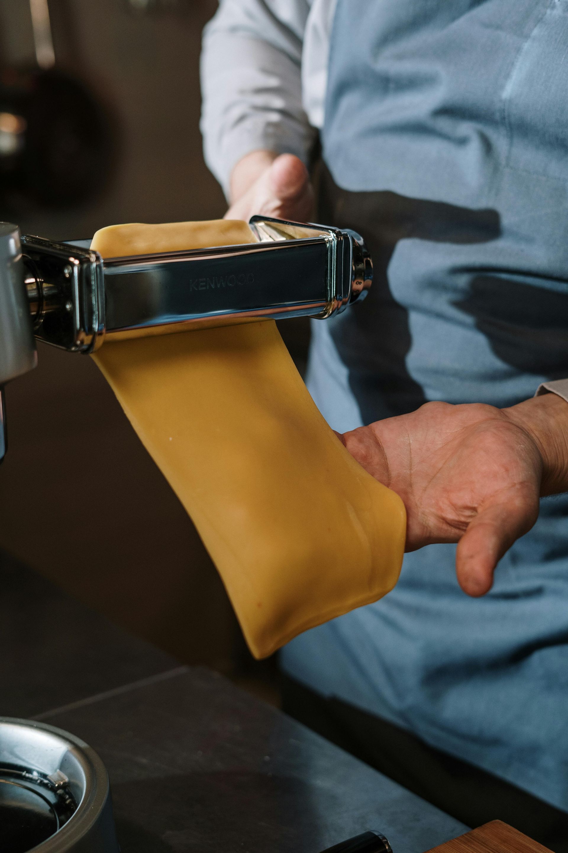 Persona che stende l'impasto della pasta fresca nella macchina per la pasta, indossando un grembiule blu.